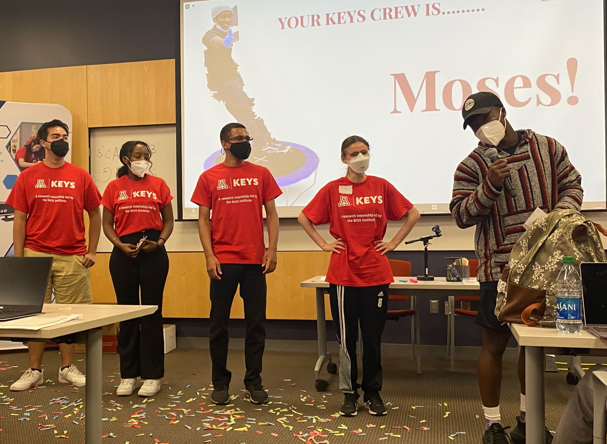 Young man with a microphone stands in front of a room with several students wearing red KEYS Research Internship shirts. All are wearing masks.