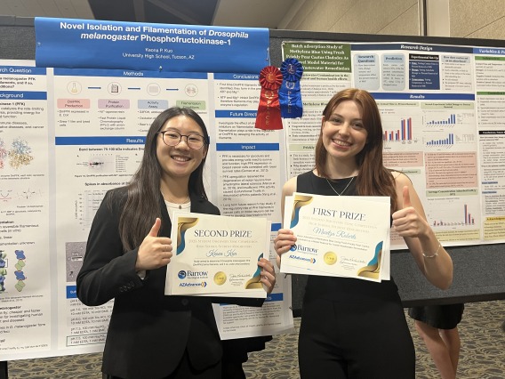 Two young women stand in front of scientific posters and give a thumbs up while showing off their awards