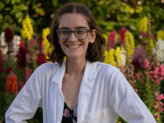 Young person wearing a lab coat smiles with her hands on her hips. Behind her are color spring flowers and greenery.