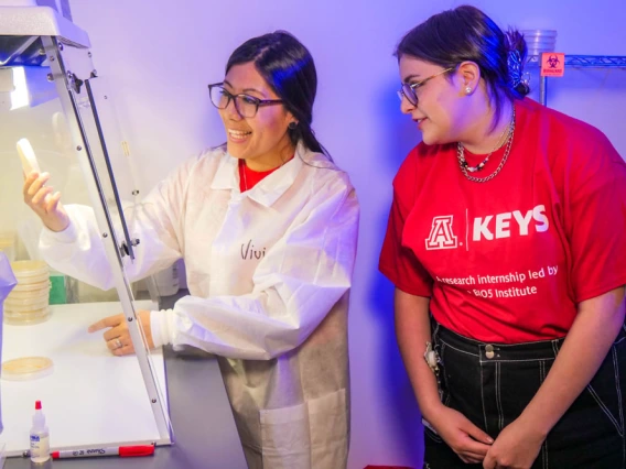 Person in white lab coat explains research to student in red KEYS t-shirt