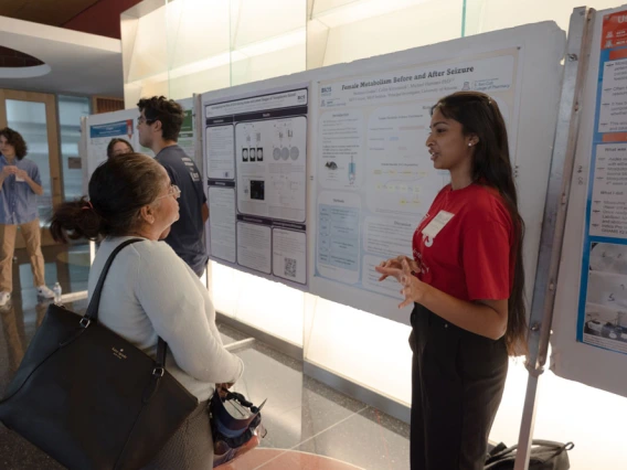 Two individuals engage in a discussion beside a research poster. One person, standing, gestures towards the poster, while the other listens attentively. Several other attendees can be seen in the background.