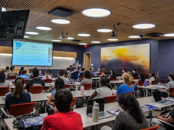 A lecture at a university classroom with students sitting at desks watching a presentation on a screen at the front. The room features modern decor with colorful wall panels and a large abstract painting on the side wall.