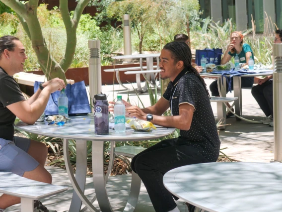 Two people engaged in conversation at an outdoor table, surrounded by lush greenery. They are seated with snacks and drinks on the table between them.