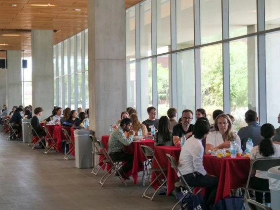 A lively scene a with individuals seated at long tables, dining and conversing, enclosed by floor-to-ceiling windows allowing ample natural light.
