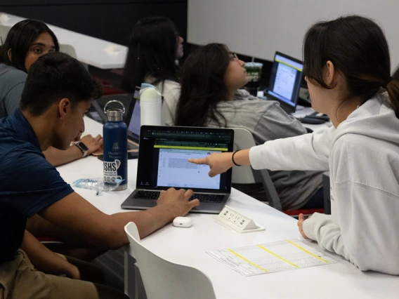 A group of students collaborate around a laptop in a classroom setting, discussing content displayed on the screen.