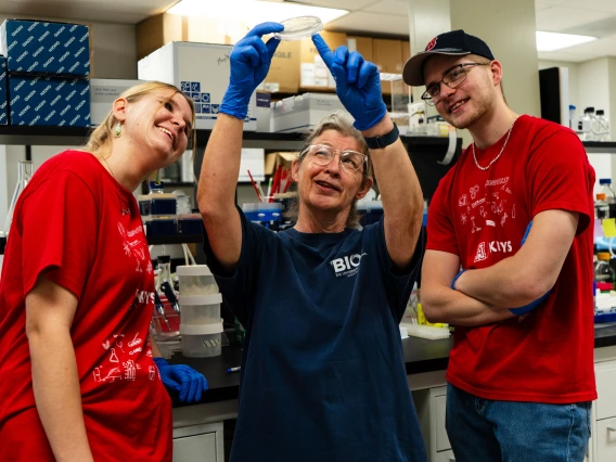 Three people in a lab, one holding a petri dish, smiling and looking at it, surrounded by lab equipment.