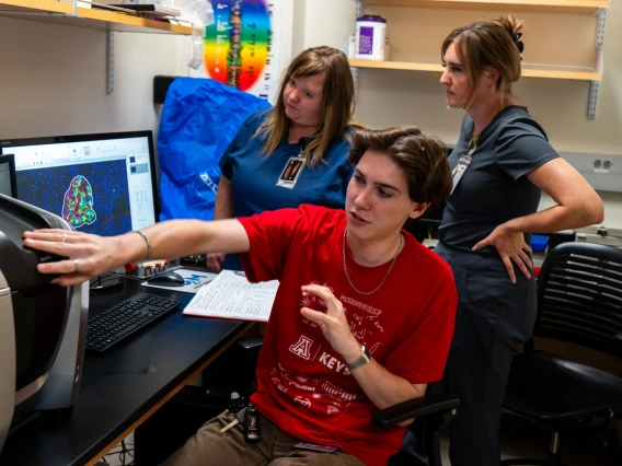 A person in a red KEYS shirt points at a computer screen displaying colorful imaging of a pancreas, while two people observe in a lab setting.