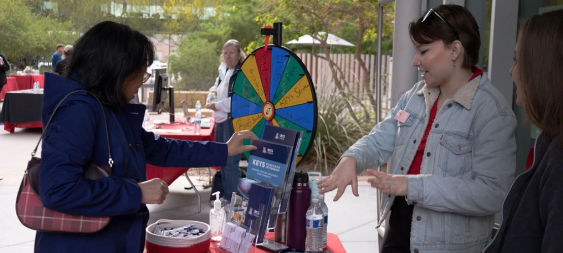 Two individuals interact at a promotional booth with a prize wheel and various branded giveaways. A third person observes nearby.