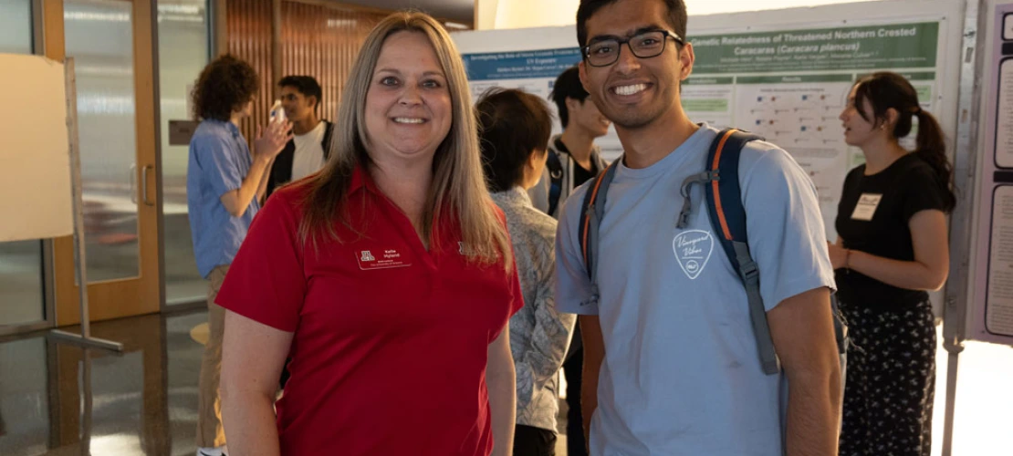 Two people smiling at a university poster presentation, one wearing a red polo with a logo and the other in a blue t-shirt with a backpack.