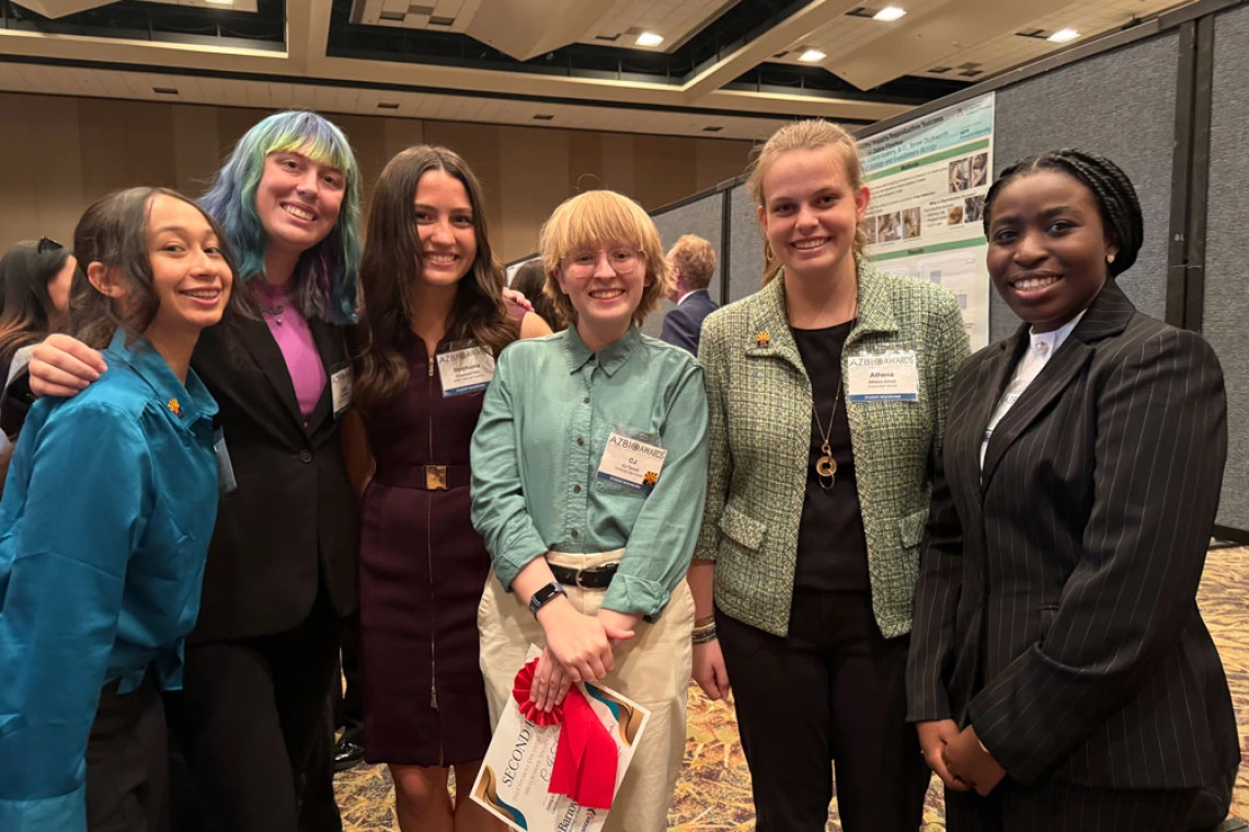 Group of six individuals smiling at a conference, standing in front of a poster presentation. They are dressed in professional attire.