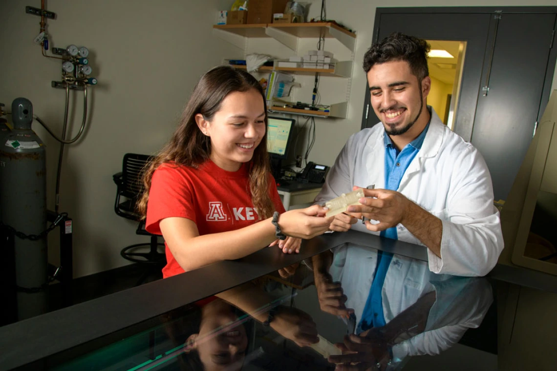 Two University of Arizona researchers in a lab, sharing and examining a 3D printed device, with scientific equipment in the background.