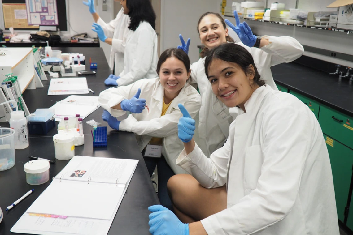 Four individuals in lab coats and gloves giving thumbs up in a laboratory setting, surrounded by scientific equipment and notebooks.