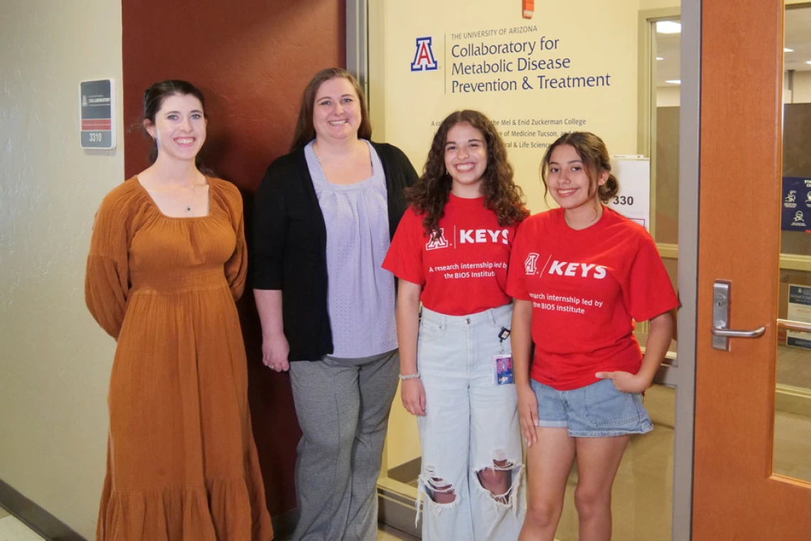 Four individuals are standing in front of the entrance to the "University of Arizona Collaboratory for Metabolic Disease Prevention & Treatment." They are smiling and dressed casually. Two of them are wearing red T-shirts with the "KEYS" logo on it.