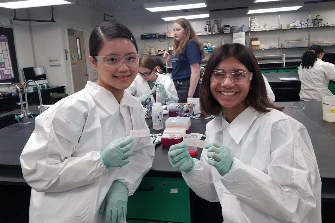 Two young people in lab white coats and safety glasses holding test tubes in a lab setting, smiling at the camera.
