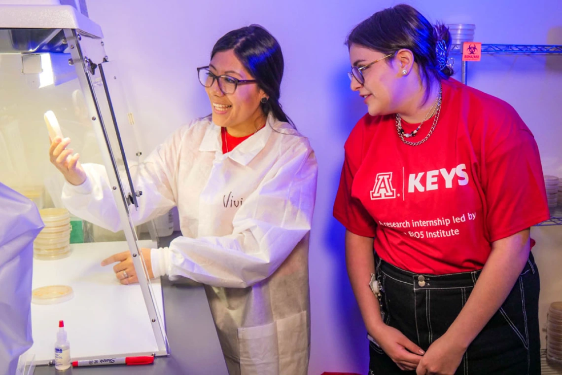 Person in white lab coat explains research to student in red KEYS t-shirt