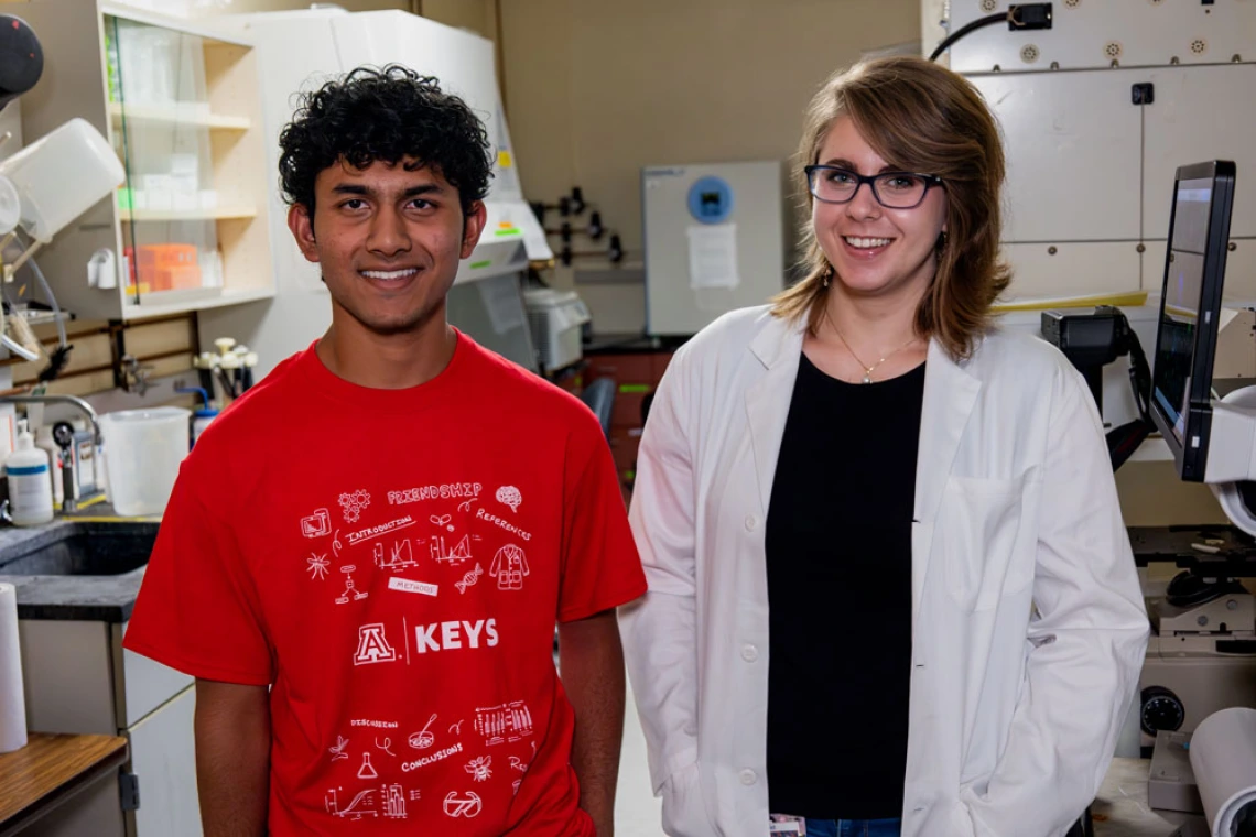 Two scientists in a laboratory, one wearing a red T-shirt and the other in a white lab coat, standing in front of complex equipment.