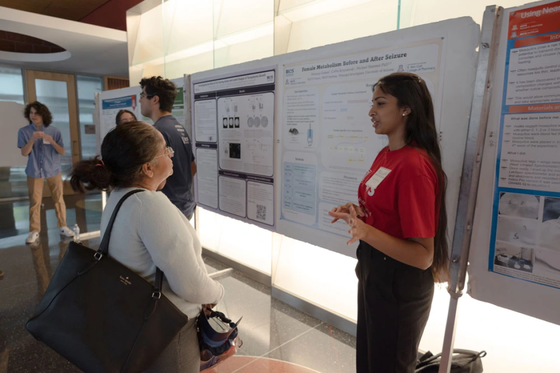 Two individuals engage in a discussion beside a research poster. One person, standing, gestures towards the poster, while the other listens attentively. Several other attendees can be seen in the background.