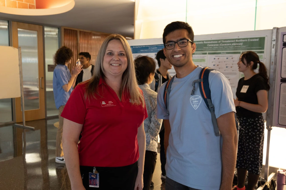 Two people smiling at a university poster presentation, one wearing a red polo with a logo and the other in a blue t-shirt with a backpack.