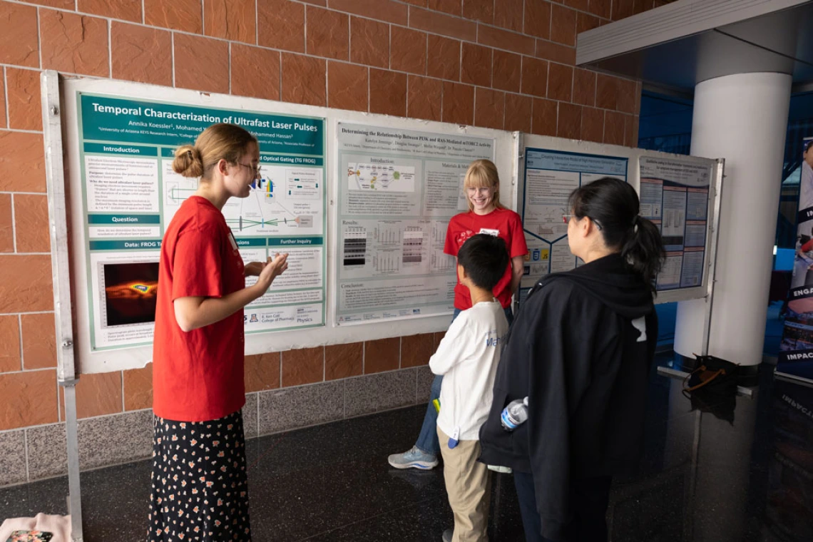 Four individuals discussing scientific posters at a conference. One person is presenting, wearing a red shirt and standing beside posters. Three attendees are listening, including a child. They are indoors with a tile floor and brick wall in the background.