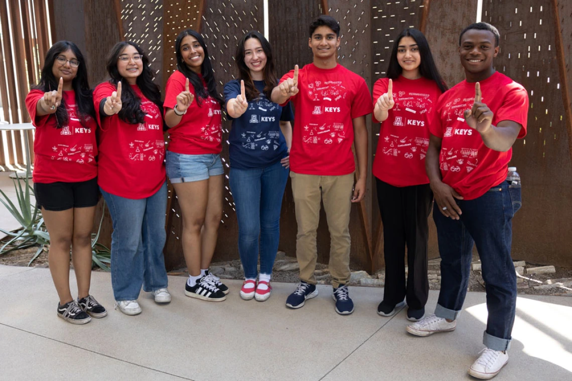 Group of six smiling students in red KEYS t-shirts pointing towards the camera, standing outdoors.