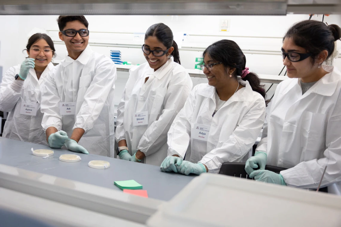 Group of five students in lab coats and gloves smiling while conducting an experiment in a laboratory.