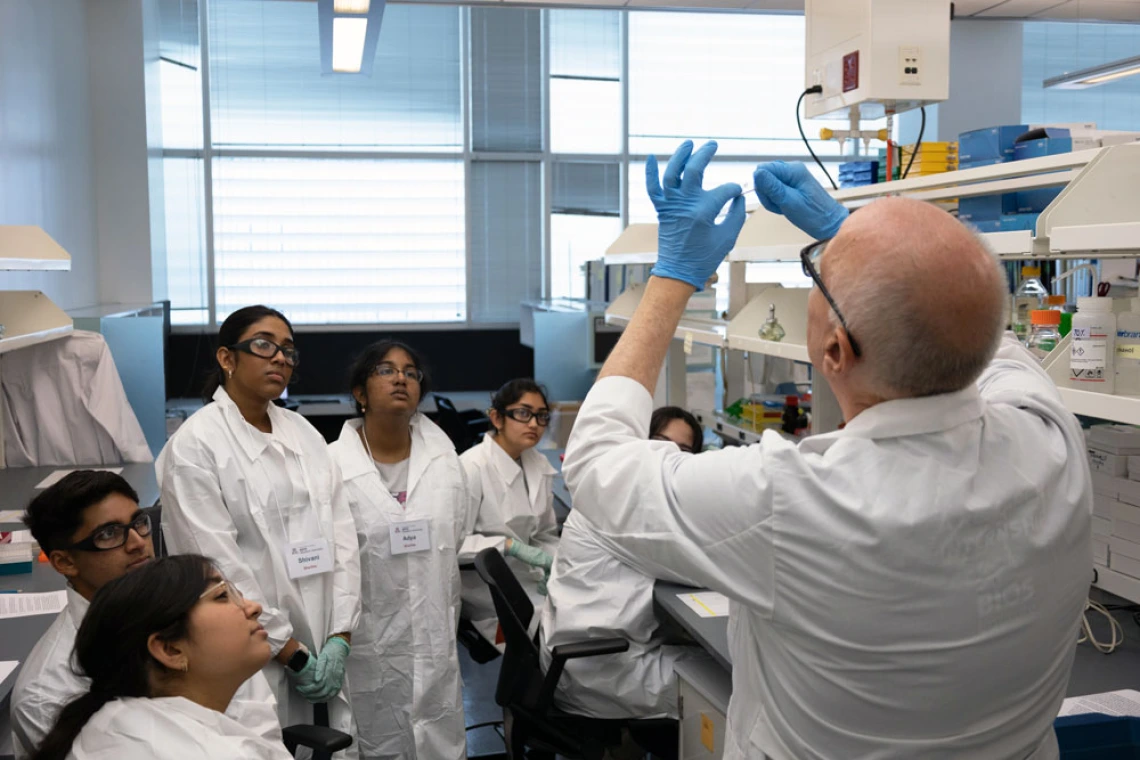 A group of students in lab coats attentively watching an instructor demonstrating a procedure in a laboratory setting.
