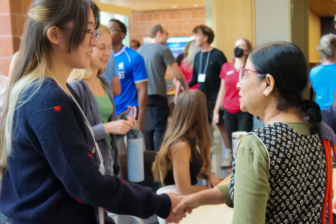 Two individuals are shaking hands at a crowded event, while engaging in conversation.