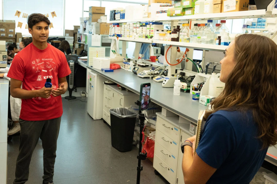 Two individuals in a laboratory setting, one in a red "KEYS" tshirt engaging in a conversation with another person in a lab filled with scientific equipment.