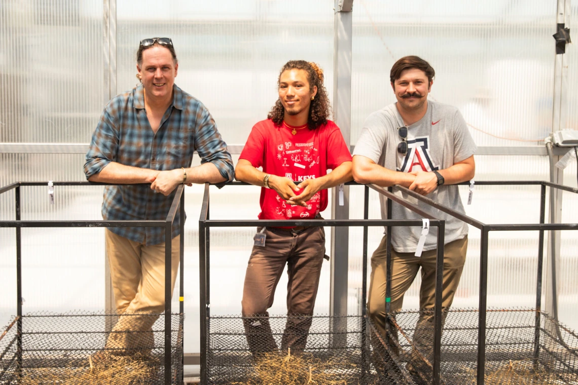 Three individuals leaning on a railing inside a greenhouse, with one wearing a University of Arizona shirt.