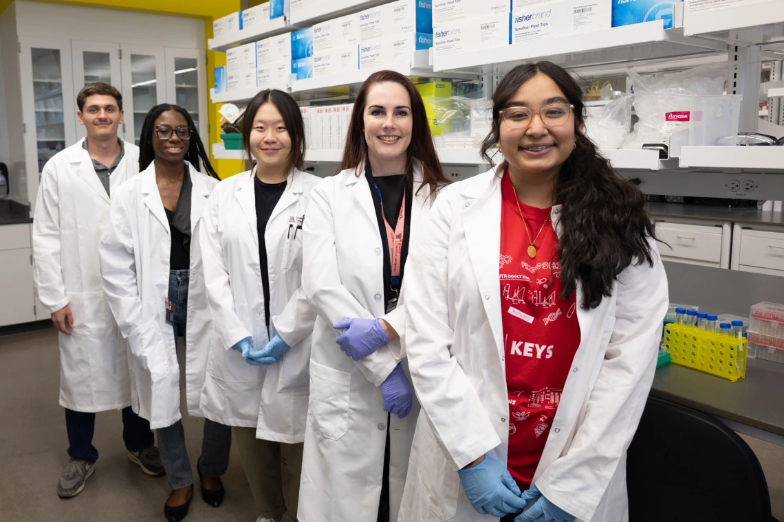Group of five diverse scientists in lab coats standing in a laboratory, each smiling at the camera.