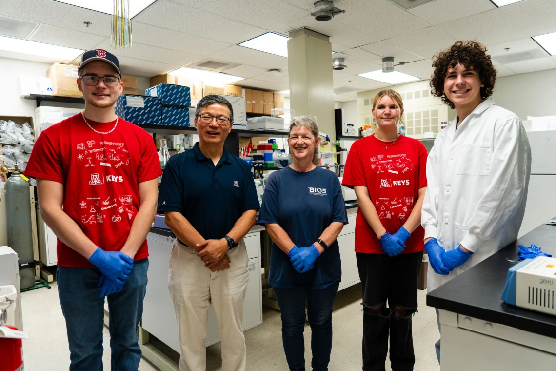 Five laboratory staff, including two wearing red shirts with "KEYS" logos, stand together in a lab setting, smiling at the camera.