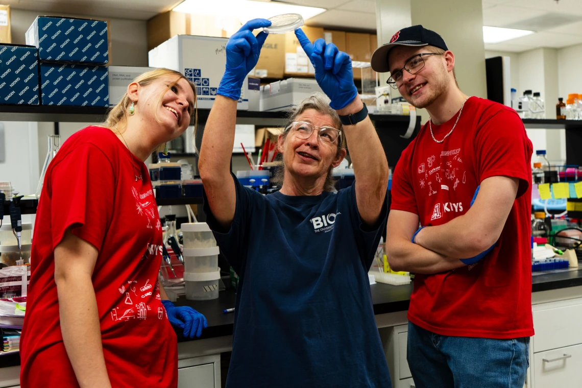 Three people in a lab, one holding a petri dish, smiling and looking at it, surrounded by lab equipment.
