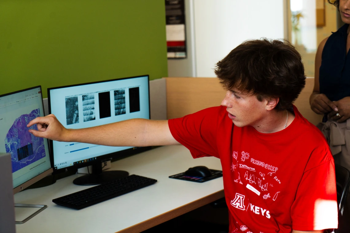 Person in a red University of Arizona T-shirt pointing at a computer screen displaying colorful graphs, with another person observing, in an office environment.