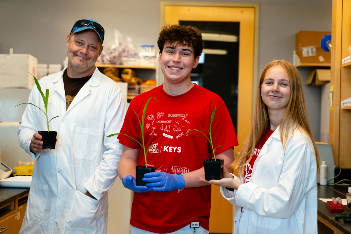 Three people in a laboratory, each holding a potted corn plant. The person in the center is wearing a red t-shirt with a "KEYS" logo, while the others wear white lab coats. They are smiling at the camera with a workbench and equipment in the background.