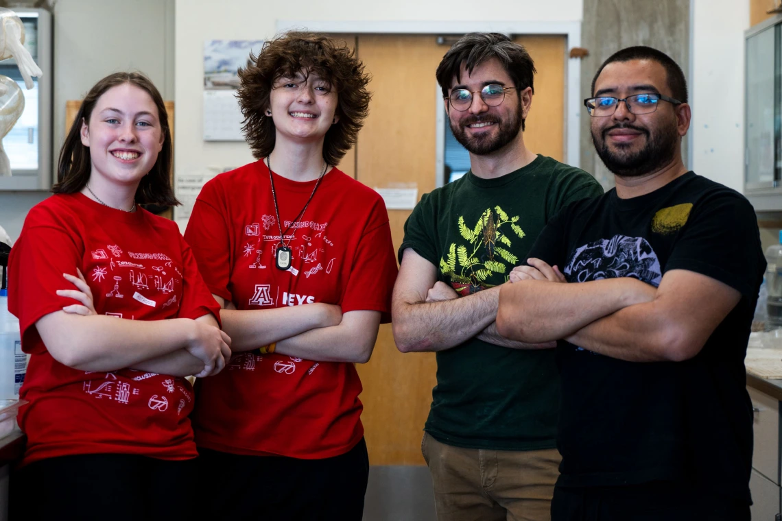 Four individuals standing side by side in a laboratory, smiling at the camera. Two are wearing red T-shirts with "KEYS" printed on them.