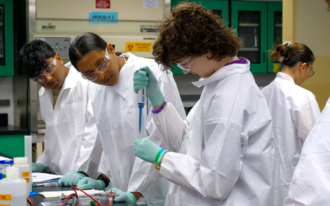 Three people in white coats are focused on conducting an experiment in a lab setting, handling test tubes and other scientific equipment.