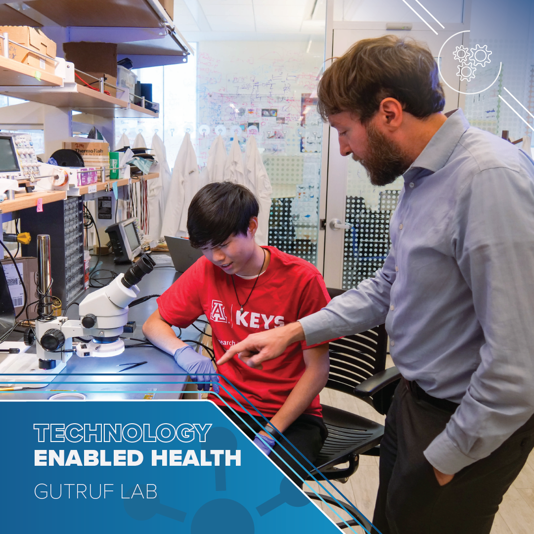 Man in a blue shirt points at a biomedical device on a lab bench. A young man wearing a red t-shirt with the KEYS logo looks on. Text reads Technology-enabled health Gutruf Lab