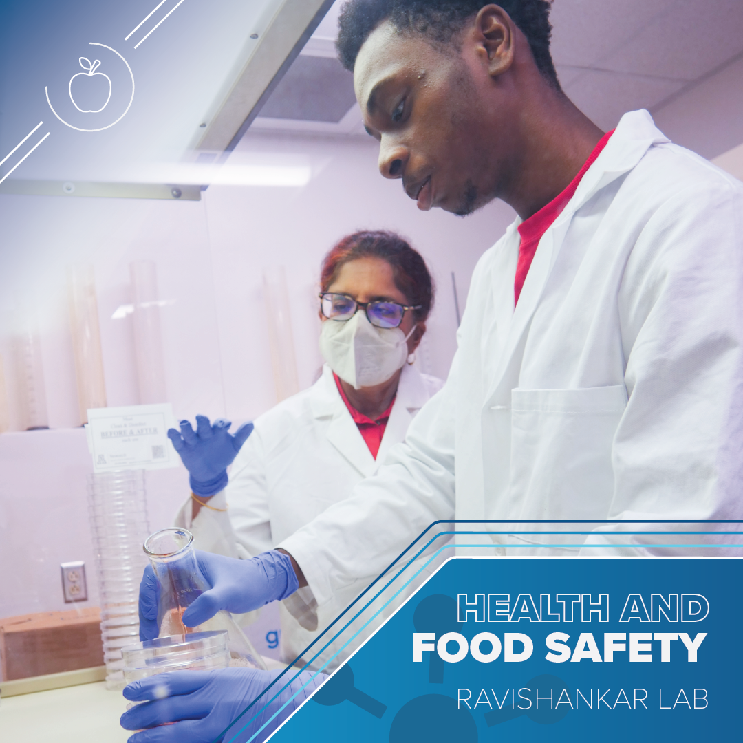 Young man in a lab coat places a vial in a  machine on a lab bench. Another older man wearing a lab coat supervises behind him. Text reads Health and Food Safety Ravishankar Lab