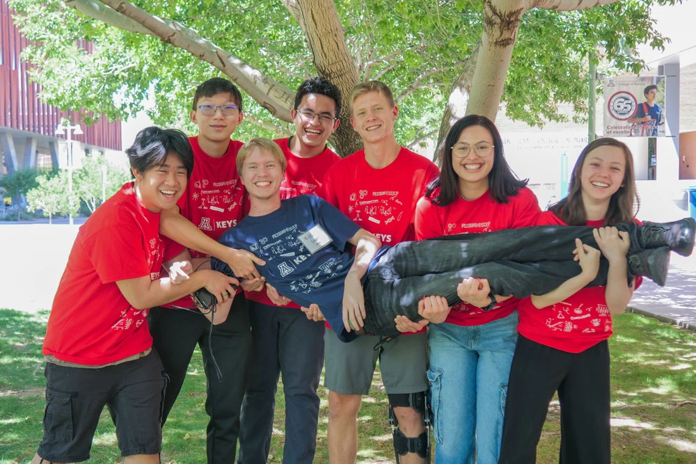 Group of seven individuals wearing red shirts, smiling and holding an individual in a blue shirt, gathered outdoors under a tree.