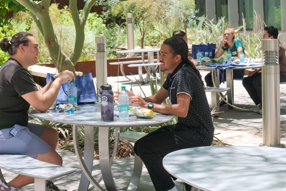 Two people engaged in conversation at an outdoor table, surrounded by lush greenery. They are seated with snacks and drinks on the table between them.