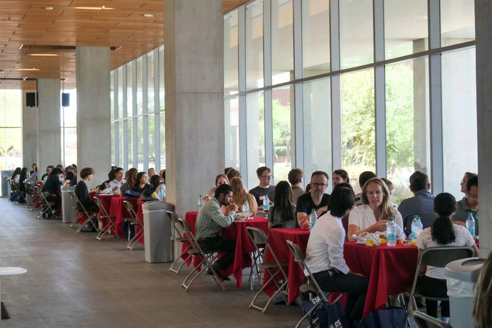 A lively scene a with individuals seated at long tables, dining and conversing, enclosed by floor-to-ceiling windows allowing ample natural light.