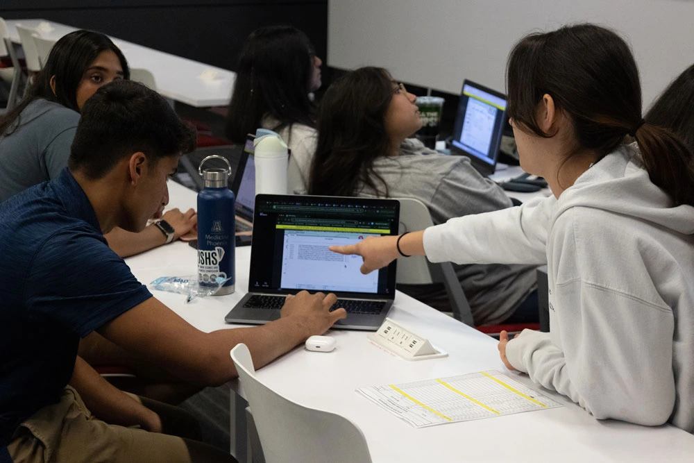 A group of students collaborate around a laptop in a classroom setting, discussing content displayed on the screen.