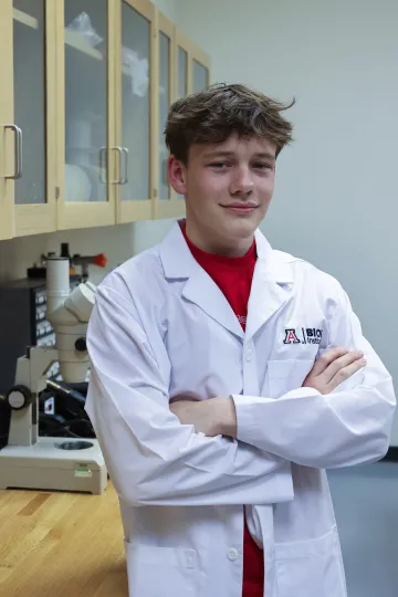 Student in a white lab coat stands confidently with crossed arms in a laboratory setting, smiling at the camera, with a microscope and other scientific equipment in the background.