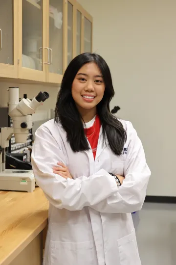 Student in a white lab coat stands confidently with crossed arms in a laboratory setting, smiling at the camera, with a microscope and other scientific equipment in the background.