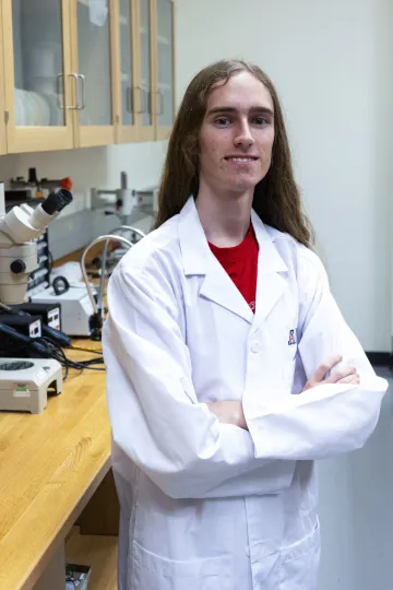 Student in a white lab coat stands confidently with crossed arms in a laboratory setting, smiling at the camera, with a microscope and other scientific equipment in the background.