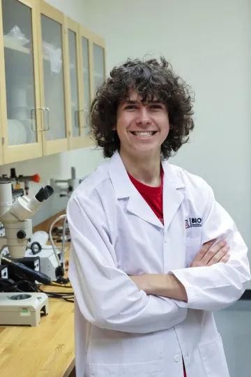 Student in a white lab coat stands confidently with crossed arms in a laboratory setting, smiling at the camera, with a microscope and other scientific equipment in the background.