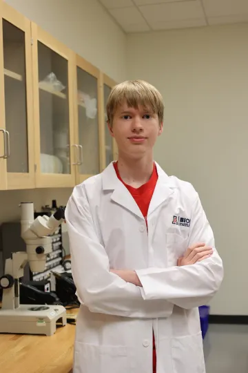 Student in a white lab coat stands confidently with crossed arms in a laboratory setting, smiling at the camera, with a microscope and other scientific equipment in the background.