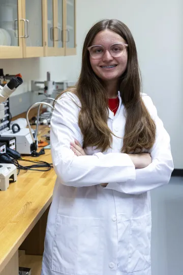 Student in a white lab coat stands confidently with crossed arms in a laboratory setting, smiling at the camera, with a microscope and other scientific equipment in the background.