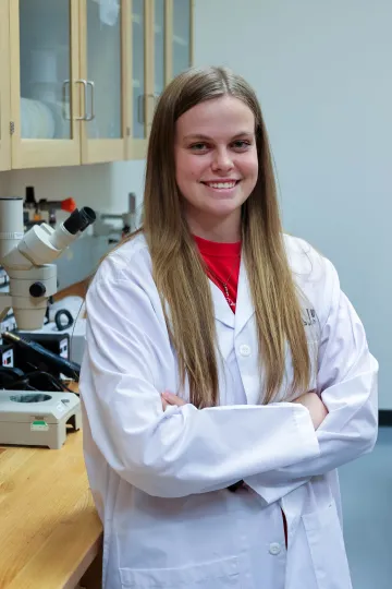 Student in a white lab coat stands confidently with crossed arms in a laboratory setting, smiling at the camera, with a microscope and other scientific equipment in the background.