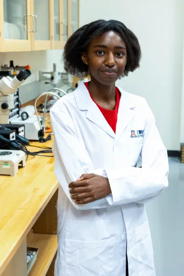 Student in a white lab coat stands confidently with crossed arms in a laboratory setting, smiling at the camera, with a microscope and other scientific equipment in the background.